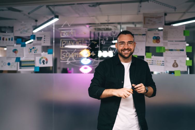 Cheerful businessman standing in office against glass wall reflecting glowing lights