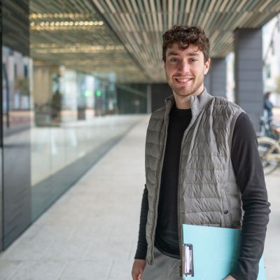 Young male professional is smiling outside office building while holding a clipboard